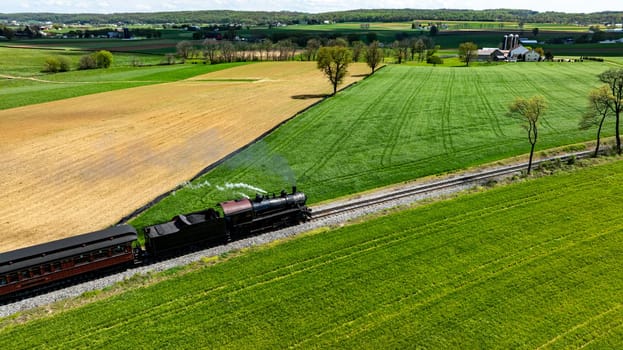 A historic steam locomotive chugs along scenic tracks, weaving through vibrant green fields and farmland under a clear blue sky. The lush landscape showcases the beauty of spring.