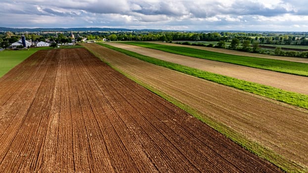 Expansive farmland stretches across the landscape, featuring alternating crop rows in varying shades of green and brown. Cloudy skies hover above the rich agricultural fields.