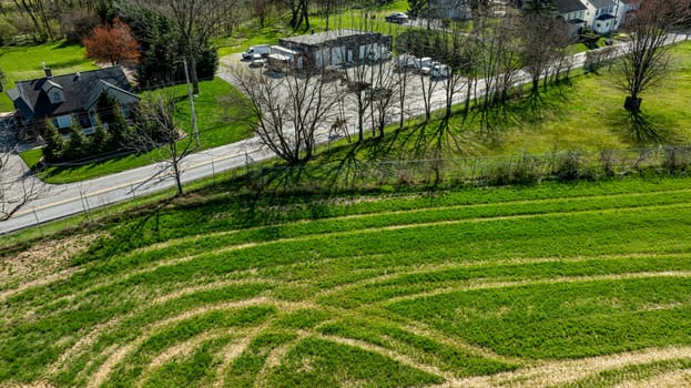 The scene captures an aerial perspective of a vibrant green field marked by gentle curves, adjacent to a winding road. Nearby, a blend of homes and vehicles adds to the rural charm of the area.