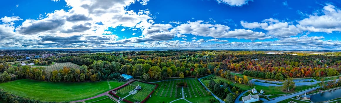 The vibrant hues of autumn foliage stretch across rolling hills under a bright blue sky scattered with fluffy clouds. The landscape showcases both trees and manicured lawns.