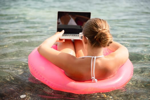 A woman is sitting on a red inflatable raft with a laptop on her lap. She is wearing a white bikini and sunglasses. Concept of relaxation and leisure