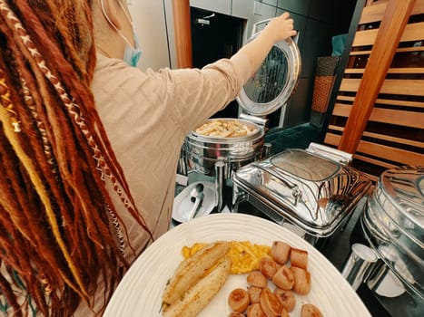 In a first-person perspective, a man is meticulously plating a dish. In a hotel, a woman is choosing food from a warming machine to fill up her plate at the buffet.