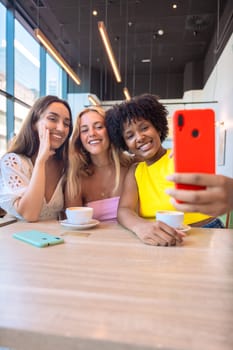 Three diverse women smiling and posing for a selfie at a cafe. They are enjoying their time together over coffee, capturing a fun moment with a smartphone. Bright, cheerful, and casual setting
