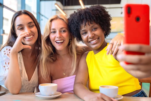 Three diverse women smiling and posing for a selfie at a cafe. They are enjoying their time together over coffee, capturing a fun moment with a smartphone. Bright, cheerful, and casual setting