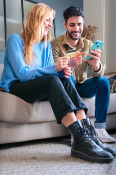 Smiling youngl couple sit at table at home using celular phone browsing wireless internet on device. A multiethnic man and woman have fun browsing the Internet on a laptop. Technology concept