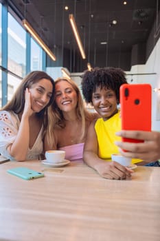 Three diverse women smiling and posing for a selfie at a cafe. They are enjoying their time together over coffee, capturing a fun moment with a smartphone. Bright, cheerful, and casual setting