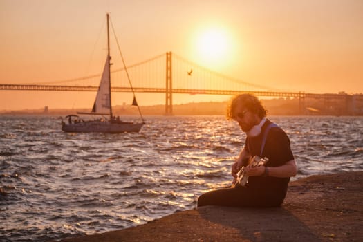Hipster street musician in black playing electric guitar in the street on sunset on embankment with 25th of April bridge and yacht boat in background. Lisbon, Portugal