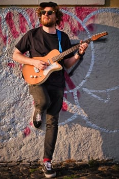 Hipster street musician in black playing electric guitar in the street on sunset leaning on a wall. Lisbon, Portugal