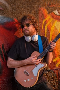 Hipster street musician in black playing electric guitar in the street on sunset leaning on a painted wall