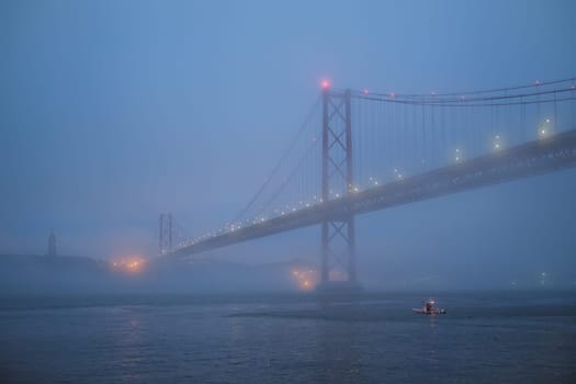 View of 25 de Abril Bridge famous tourist landmark of Lisbon connecting Lisboa and Almada in heavy fog mist wtih yacht boats passing under. Lisbon, Portugal
