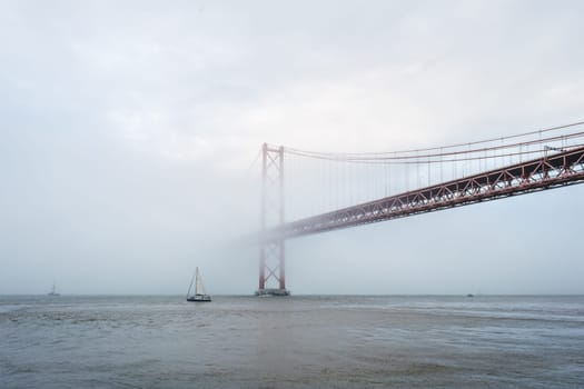 View of 25 de Abril Bridge famous tourist landmark of Lisbon connecting Lisboa and Almada in heavy fog mist wtih yacht boats passing under. Lisbon, Portugal