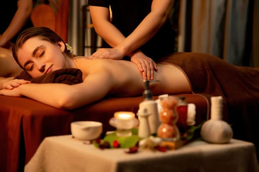 Caucasian couple customer enjoying relaxing anti-stress spa massage and pampering with beauty skin recreation leisure in warm candle lighting ambient salon spa at luxury resort or hotel. Quiescent