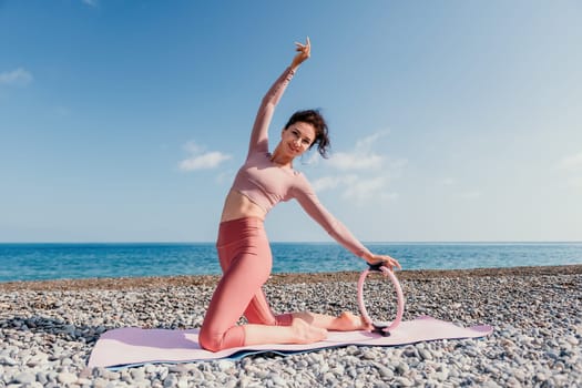 Middle aged well looking woman with black hair doing Pilates with the ring on the yoga mat near the sea on the pebble beach. Female fitness yoga concept. Healthy lifestyle, harmony and meditation.