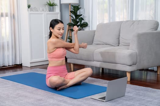 Asian woman in sportswear doing yoga exercise on fitness mat as her home workout training routine. Healthy body care lifestyle woman watching online yoga video on laptop. Vigorous