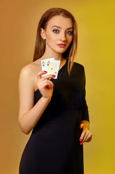 Beautiful blonde model with bright make-up, red manicure, in black dress is showing two aces, hand on hip, posing against colorful background. Gambling entertainment, poker, casino. Close-up.
