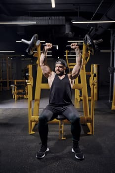 Tattooed, bearded male in black sweatpants, vest and cap. He performing a chest press while sitting on an exercise machine at dark gym with yellow equipment. Sport, fitness, lifestyle. Full length