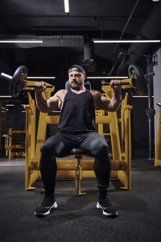 Tattooed, bearded, strong guy in black sweatpants, vest and cap. He performing a chest press while sitting on an exercise machine at dark gym with yellow equipment. Sport, fitness. Full length