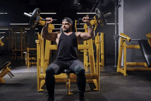 Tattooed, bearded sportsman in black sweatpants, vest and cap. He performing a chest press while sitting on an exercise machine at dark gym with yellow equipment. Sport, fitness. Close up