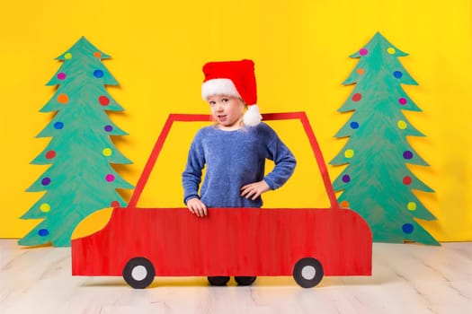 Child with Christmas hat driving a car made of cardboard. Little girl having fun at home on a yellow background. Christmas concept. New Year's holidays.