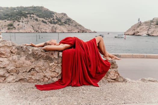 Side view a Young beautiful sensual woman in a red long dress posing on a volcanic rock high above the sea during sunset. Girl on the nature on overcast sky background. Fashion photo