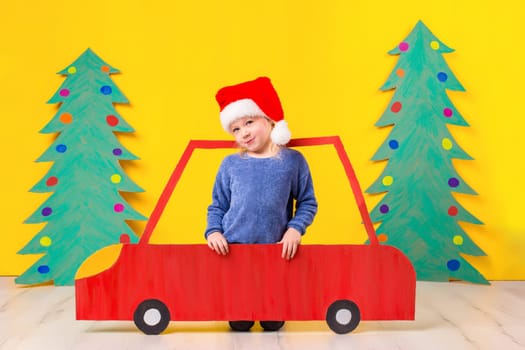 Child with Christmas hat driving a car made of cardboard. Little girl having fun at home on a yellow background. Christmas concept. New Year's holidays.