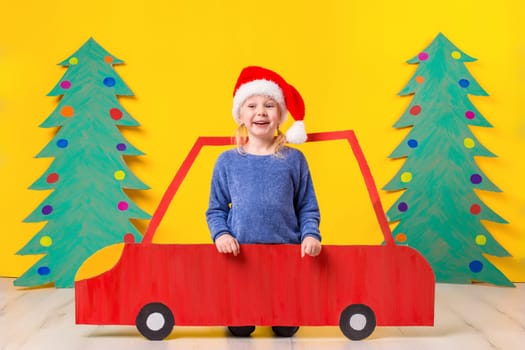 Child with Christmas hat driving a car made of cardboard. Little girl having fun at home on a yellow background. Christmas concept. New Year's holidays.