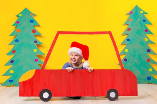 Child with Christmas hat driving a car made of cardboard. Little girl having fun at home on a yellow background. Christmas concept. New Year's holidays.