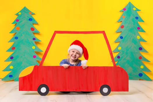 Child with Christmas hat driving a car made of cardboard. Little girl having fun at home on a yellow background. Christmas concept. New Year's holidays.