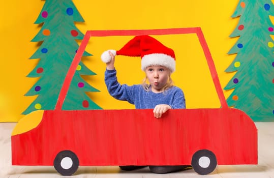 Child with Christmas hat driving a car made of cardboard. Little girl having fun at home on a yellow background. Christmas concept. New Year's holidays.