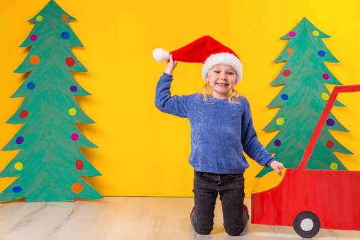 Child with Christmas hat driving a car made of cardboard. Little girl having fun at home on a yellow background. Christmas concept. New Year's holidays.