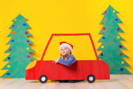 Child in red Christmas car. A painted car and green Christmas tree made of cardboard on a yellow background. Xmas holiday concept. Little emotional girl