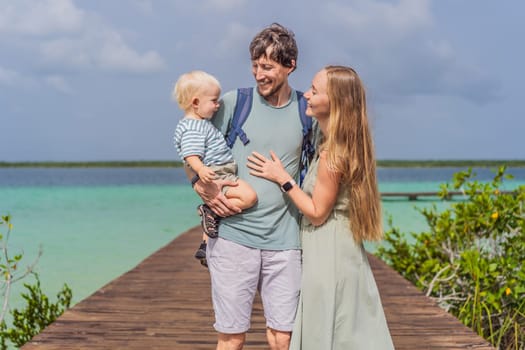 Mom, dad and their baby son exploring wooden pier over the turquoise waters of Bacalar Lake, Mexico. Peaceful tropical travel destination concept.