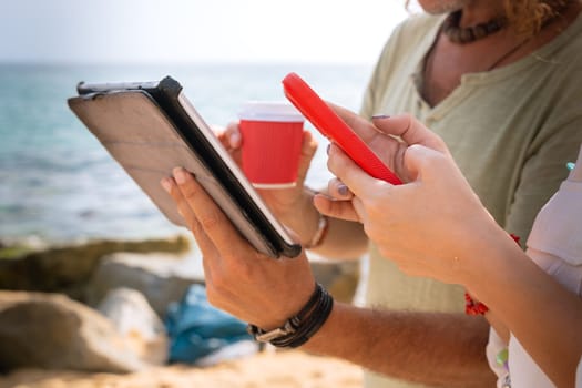 Middle-aged man and woman sitting on the beach with surprised face, browsing smartphone and tablet apps. Concept: Holidays and Technology