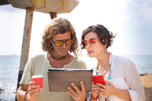 Middle-aged man and woman sitting on the beach with surprised face, browsing smartphone and tablet apps. Concept: Holidays and Technology