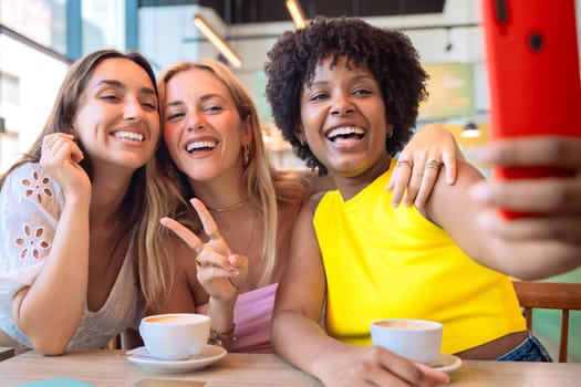 Three diverse women smiling and posing for a selfie at a cafe. They are enjoying their time together over coffee, capturing a fun moment with a smartphone. Bright, cheerful, and casual setting