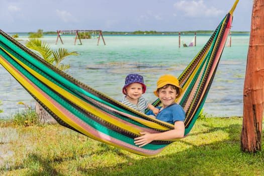 Two boys, a baby and a teenager lying in a hammock against the backdrop of turquoise waters of Bacalar Lake, Mexico. Peaceful tropical travel destination concept.