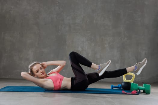 A woman in athletic wear performing bicycle crunches on a blue exercise mat indoors. Various fitness equipment is placed nearby.