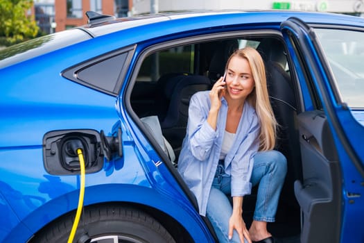 Woman is seated in her electric car, charging it and talking on her smartphone outdoors.