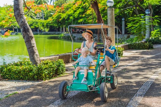 Mother tourist with her toddler and teenage sons riding an authentic Mexican pedal car in a park in Mexico. Family adventure and travel with kids concept.