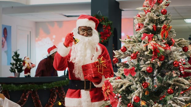 African american employee posing as Santa Claus ringing bells next to beautifully decorated Christmas tree, trying to attract customers to fashion store during promotional holiday season