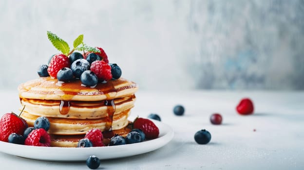 A beautiful stack of fluffy pancakes sits on a white plate, generously topped with an assortment of fresh blueberries and raspberries. The pancakes are drizzled with golden maple syrup and garnished with a sprig of mint, creating an inviting and delicious breakfast treat. The light, airy background enhances the vibrant colors of the berries and syrup.