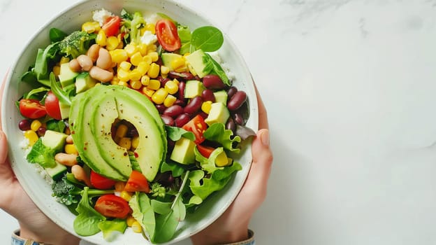 A person holds a large bowl of vibrant salad filled with greens, tomatoes, corn, and beans. The dish features beautifully sliced avocado on top, adding a creamy texture. The setting is bright and inviting, perfect for a healthy meal.