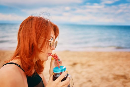 Red-haired woman with sunglasses on vacation. Holidays at the sea. Calm lady in a swimsuit is sitting on beach with a cocktail and look away. Tropical alcohol beverage. Summer day with cloudy sky.