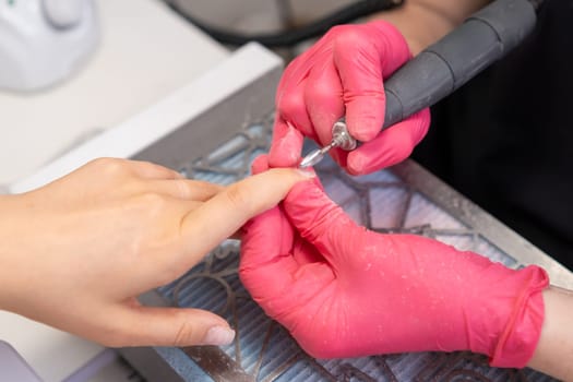 Nail master in pink gloves remove old gel polish using nail dril machine in beauty salon.