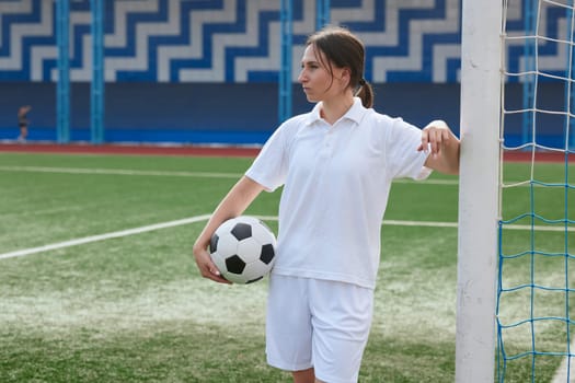 Female soccer player in white uniform holding a soccer ball and leaning against a goalpost on a soccer field.
