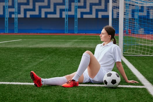A female football player sits on the grass of a football field, wearing a white T-shirt and shorts, with red football boots. break after a football match