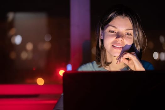 A young girl works at home at night with a laptop online, a woman with a smiley face is studying, looking, searching for information, using her device