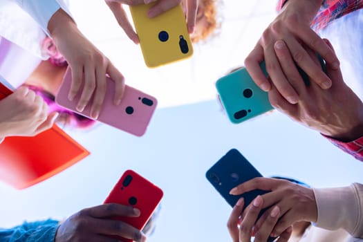 Low-angle shot of a group of diverse hands holding brightly colored smartphones, forming a circle. The photo showcases modern communication and social media interaction in a vibrant, connected setting