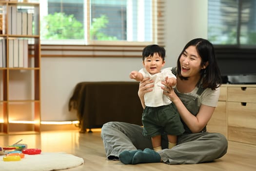 Happy young mother playing cute little boy on floor at home. Childhood and nurturing care concept.