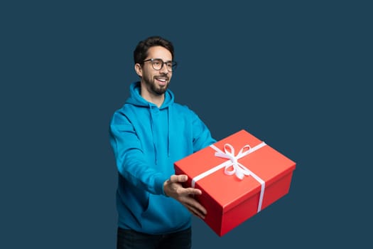 A cheerful young man in a blue hoodie extends his arms to present a large red gift box, which is tied with a white ribbon, showcasing a festive atmosphere.